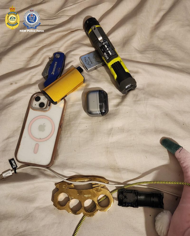 Knuckle dusters, a folding knife, lighter, torch and mobile phone laid out on a bed sheet