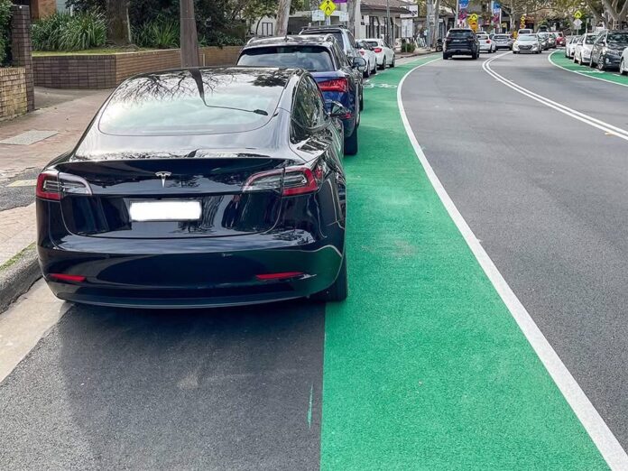 Parked cars line the kerb beside a bright green cycle lane on a Lane Cove shopping street