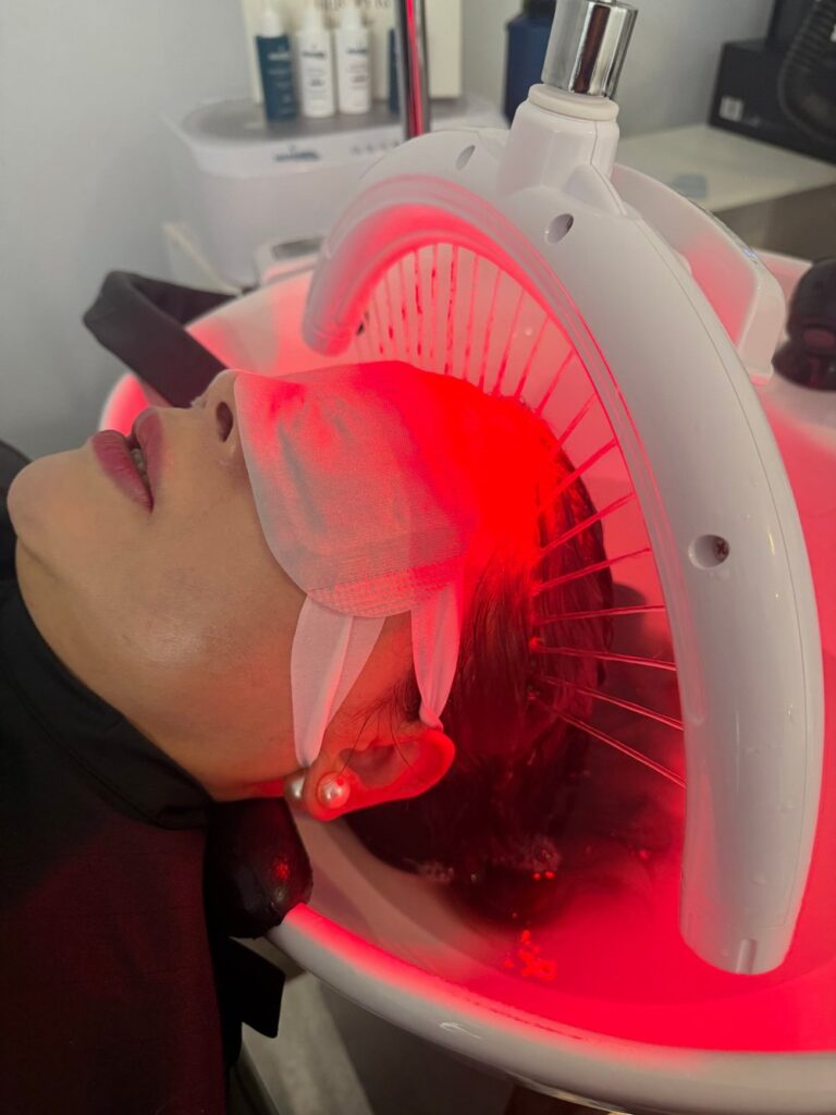 A client wearing an eye mask lies under the water arc and red light of the head spa at K Avenue Hair & Beauty.